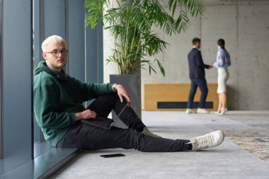 Modern bir ofiste geniş bir pencerenin yanında oturan sıradan bir profesyonel bilgisayarla çalışırken gülümseyen meslektaşları bulanık arka planda etkileşime giren biri.