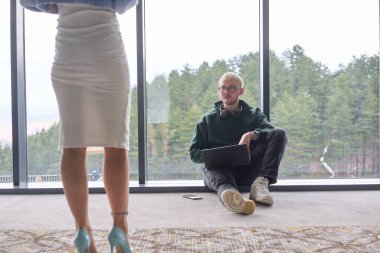 Sarışın bir adam modern bir ofisin zemininde rastgele oturuyor, dizüstü bilgisayarını kullanırken pencereye yaslanıyor, yanında çekici bayan arkadaşı duruyor..