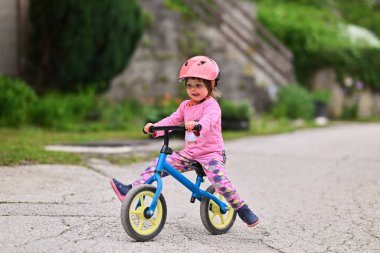 Şirin bir kostüm giymiş ve kask takan küçük bir kız heyecanlı bir şekilde ilk kez bisiklet sürmeyi öğreniyor, neşe ve kararlılıkla dolu.