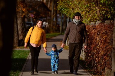 A loving couple strolls through a sunlit park with their young son, surrounded by the vibrant colors of autumn, enjoying a joyful and peaceful family moment together clipart