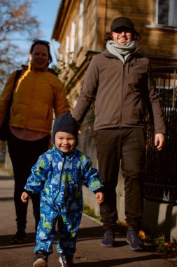 A loving couple strolls through a sunlit park with their young son, surrounded by the vibrant colors of autumn, enjoying a joyful and peaceful family moment together clipart