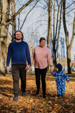 A loving couple strolls through a sunlit park with their young son, surrounded by the vibrant colors of autumn, enjoying a joyful and peaceful family moment together clipart