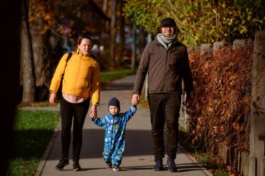 A loving couple strolls through a sunlit park with their young son, surrounded by the vibrant colors of autumn, enjoying a joyful and peaceful family moment together clipart