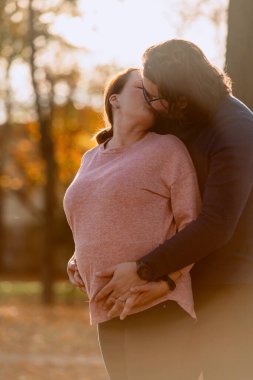 A man lovingly embraces his pregnant wife in the park, sharing a peaceful moment of warmth and affection. clipart