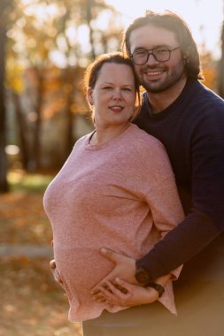 Bir adam parkta hamile karısını sevgiyle kucaklıyor. Huzurlu bir sıcaklık ve şefkat anı yaşıyor..