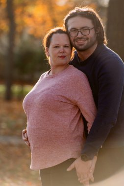 A man lovingly embraces his pregnant wife in the park, sharing a peaceful moment of warmth and affection. clipart