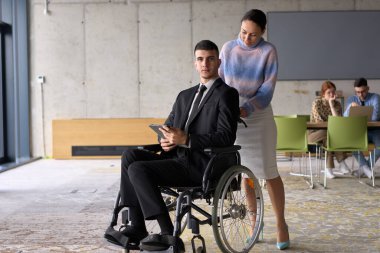 In a professional office setting, a businesswoman helping her director, who is sitting in a wheelchair and using a tablet, while their colleagues collaborate in the background clipart