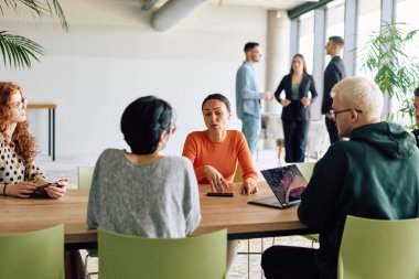 A diverse team of business professionals engaged in a discussion around a conference table in an office, while their colleagues collaborate in the background.  clipart