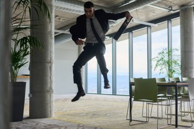 Modern bir ofise kendinden geçmiş bir şekilde atlayan bir işadamı, elinde bir evrak çantasıyla, başarı ve zafer anını kutluyor.