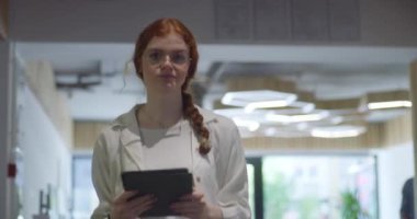 Canlı turuncu saçlı modern bir iş kadını, profesyonelliği, dijital zekayı ve verimliliği temsil eden çağdaş bir ofis alanında gezinirken kendine güvenen bir tablet tutuyor.