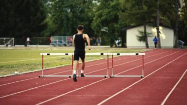 Güçlü Erkek Sporcu bir engele doğru koşuyor, bir yarışta koşarken bariyerin üzerinden yüksek hızda atlıyor. Hız Rampası Efektli Süper Yavaş Sinema Görüntüsü.