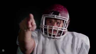 Takım üniformalı, pedli ve kasklı Amerikalı futbolcu kameraya doğru bakıyor. Güçlü, aksiyon dolu, takım ruhunu yakalayan ve oyun için hazır olan bir görüntü. Sinematik ağır çekim