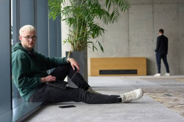 Modern bir ofiste geniş bir pencerenin yanında oturan sıradan bir profesyonel bilgisayarla çalışırken gülümseyen meslektaşları bulanık arka planda etkileşime giren biri.