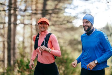 Gülümseyen bir erkek ve kadın, serin bir günde, doğa, fitness ve birbirlerine eşlik ederek, spor giysileri içinde, manzaralı bir orman patikasında birlikte koşarlar..