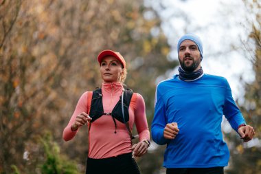 Gülümseyen bir erkek ve kadın, serin bir günde, doğa, fitness ve birbirlerine eşlik ederek, spor giysileri içinde, manzaralı bir orman patikasında birlikte koşarlar..