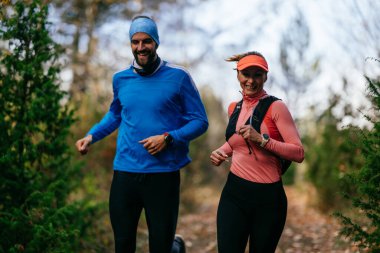 Gülümseyen bir erkek ve kadın, serin bir günde, doğa, fitness ve birbirlerine eşlik ederek, spor giysileri içinde, manzaralı bir orman patikasında birlikte koşarlar..