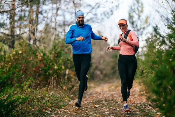 Gülümseyen bir erkek ve kadın, serin bir günde, doğa, fitness ve birbirlerine eşlik ederek, spor giysileri içinde, manzaralı bir orman patikasında birlikte koşarlar..