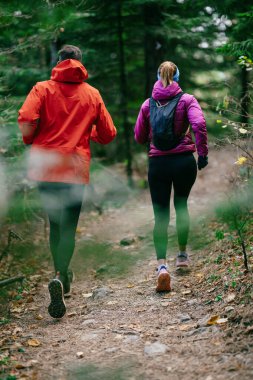 Gülümseyen bir erkek ve kadın, serin bir günde, doğa, fitness ve birbirlerine eşlik ederek, spor giysileri içinde, manzaralı bir orman patikasında birlikte koşarlar..