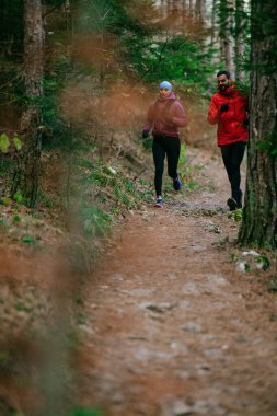 Gülümseyen bir erkek ve kadın, serin bir günde, doğa, fitness ve birbirlerine eşlik ederek, spor giysileri içinde, manzaralı bir orman patikasında birlikte koşarlar..