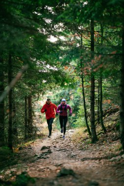 Gülümseyen bir erkek ve kadın, serin bir günde, doğa, fitness ve birbirlerine eşlik ederek, spor giysileri içinde, manzaralı bir orman patikasında birlikte koşarlar..
