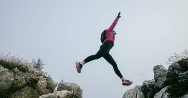 A woman athlete running along a rocky mountain trail in cold, misty weather, wearing a warm jacket, gloves, and a headband, showcasing endurance and determination.  clipart