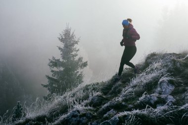 Soğuk ve sisli havada kayalık bir dağ yolunda koşan bir kadın sporcu, sıcak bir ceket, eldiven ve saç bandı giyiyor, dayanıklılık ve kararlılık sergiliyor.. 