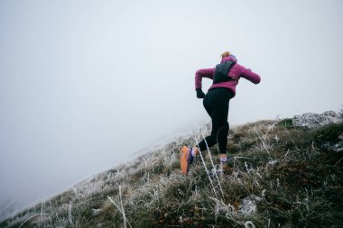 Soğuk ve sisli havada kayalık bir dağ yolunda koşan bir kadın sporcu, sıcak bir ceket, eldiven ve saç bandı giyiyor, dayanıklılık ve kararlılık sergiliyor.. 