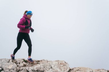 Soğuk ve sisli havada kayalık bir dağ yolunda koşan bir kadın sporcu, sıcak bir ceket, eldiven ve saç bandı giyiyor, dayanıklılık ve kararlılık sergiliyor.. 