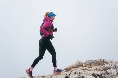 A woman athlete running along a rocky mountain trail in cold, misty weather, wearing a warm jacket, gloves, and a headband, showcasing endurance and determination.  clipart