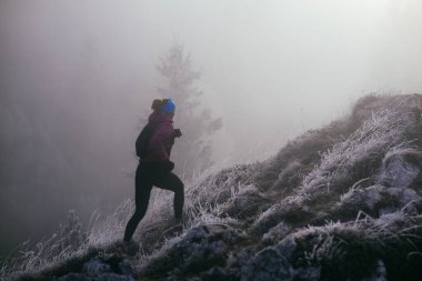 Soğuk ve sisli havada kayalık bir dağ yolunda koşan bir kadın sporcu, sıcak bir ceket, eldiven ve saç bandı giyiyor, dayanıklılık ve kararlılık sergiliyor.. 