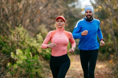 Gülümseyen bir erkek ve kadın, serin bir günde, doğa, fitness ve birbirlerine eşlik ederek, spor giysileri içinde, manzaralı bir orman patikasında birlikte koşarlar..