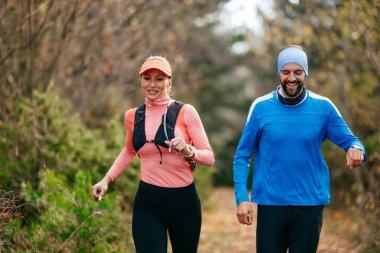 Gülümseyen bir erkek ve kadın, serin bir günde, doğa, fitness ve birbirlerine eşlik ederek, spor giysileri içinde, manzaralı bir orman patikasında birlikte koşarlar..