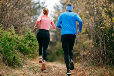 Gülümseyen bir erkek ve kadın, serin bir günde, doğa, fitness ve birbirlerine eşlik ederek, spor giysileri içinde, manzaralı bir orman patikasında birlikte koşarlar..