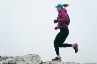 Soğuk ve sisli havada kayalık bir dağ yolunda koşan bir kadın sporcu, sıcak bir ceket, eldiven ve saç bandı giyiyor, dayanıklılık ve kararlılık sergiliyor.. 