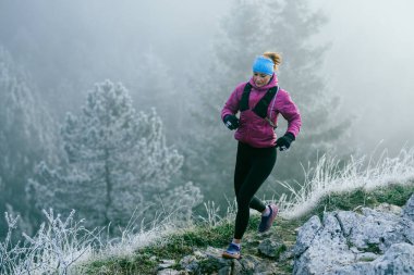 Soğuk ve sisli havada kayalık bir dağ yolunda koşan bir kadın sporcu, sıcak bir ceket, eldiven ve saç bandı giyiyor, dayanıklılık ve kararlılık sergiliyor.. 