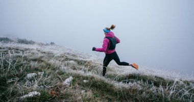 Soğuk ve sisli havada kayalık bir dağ yolunda koşan bir kadın sporcu, sıcak bir ceket, eldiven ve saç bandı giyiyor, dayanıklılık ve kararlılık sergiliyor.. 