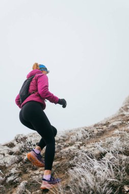 Soğuk ve sisli havada kayalık bir dağ yolunda koşan bir kadın sporcu, sıcak bir ceket, eldiven ve saç bandı giyiyor, dayanıklılık ve kararlılık sergiliyor.. 