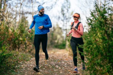 Gülümseyen bir erkek ve kadın, serin bir günde, doğa, fitness ve birbirlerine eşlik ederek, spor giysileri içinde, manzaralı bir orman patikasında birlikte koşarlar..