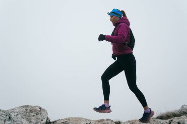Soğuk ve sisli havada kayalık bir dağ yolunda koşan bir kadın sporcu, sıcak bir ceket, eldiven ve saç bandı giyiyor, dayanıklılık ve kararlılık sergiliyor.. 