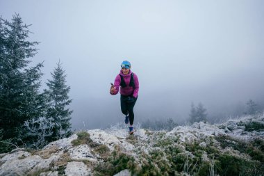 Soğuk ve sisli havada kayalık bir dağ yolunda koşan bir kadın sporcu, sıcak bir ceket, eldiven ve saç bandı giyiyor, dayanıklılık ve kararlılık sergiliyor.. 