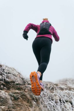 Soğuk ve sisli havada kayalık bir dağ yolunda koşan bir kadın sporcu, sıcak bir ceket, eldiven ve saç bandı giyiyor, dayanıklılık ve kararlılık sergiliyor.. 
