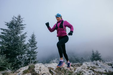 Soğuk ve sisli havada kayalık bir dağ yolunda koşan bir kadın sporcu, sıcak bir ceket, eldiven ve saç bandı giyiyor, dayanıklılık ve kararlılık sergiliyor.. 