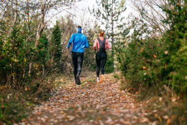 Gülümseyen bir erkek ve kadın, serin bir günde, doğa, fitness ve birbirlerine eşlik ederek, spor giysileri içinde, manzaralı bir orman patikasında birlikte koşarlar..