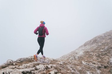 Soğuk ve sisli havada kayalık bir dağ yolunda koşan bir kadın sporcu, sıcak bir ceket, eldiven ve saç bandı giyiyor, dayanıklılık ve kararlılık sergiliyor.. 