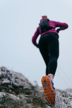 Soğuk ve sisli havada kayalık bir dağ yolunda koşan bir kadın sporcu, sıcak bir ceket, eldiven ve saç bandı giyiyor, dayanıklılık ve kararlılık sergiliyor.. 
