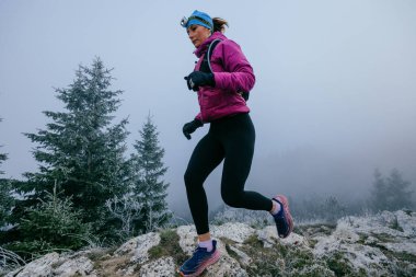 Soğuk ve sisli havada kayalık bir dağ yolunda koşan bir kadın sporcu, sıcak bir ceket, eldiven ve saç bandı giyiyor, dayanıklılık ve kararlılık sergiliyor.. 