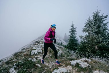 Soğuk ve sisli havada kayalık bir dağ yolunda koşan bir kadın sporcu, sıcak bir ceket, eldiven ve saç bandı giyiyor, dayanıklılık ve kararlılık sergiliyor.. 