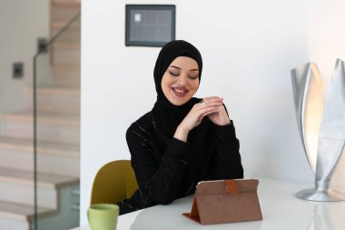 Kendine güveni tam, siyah başörtülü genç bir Müslüman kadın modern bir masada tablet, fincan ve dekoratif lambayla modern bir iç mekanda oturuyor.. 