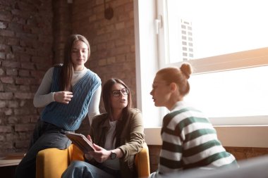 Kadın iş adamları pencere kenarında iş birliği yapıyor, fikirleri tartışıyor ve tablet üzerinde beyin fırtınası yapıyorlar. Modern bir ofiste işbirliği, yaratıcılık ve takım çalışması kavramı