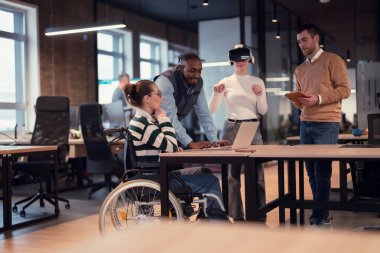 Modern bir iş yerinde çalışan tekerlekli sandalyedeki engelli bir iş kadını. Çevredeki ekip sanal gerçeklik gözlükleri kullanıyor, açık alan yaratıcılığında en son teknolojiyle işbirliği yapıyorlar.